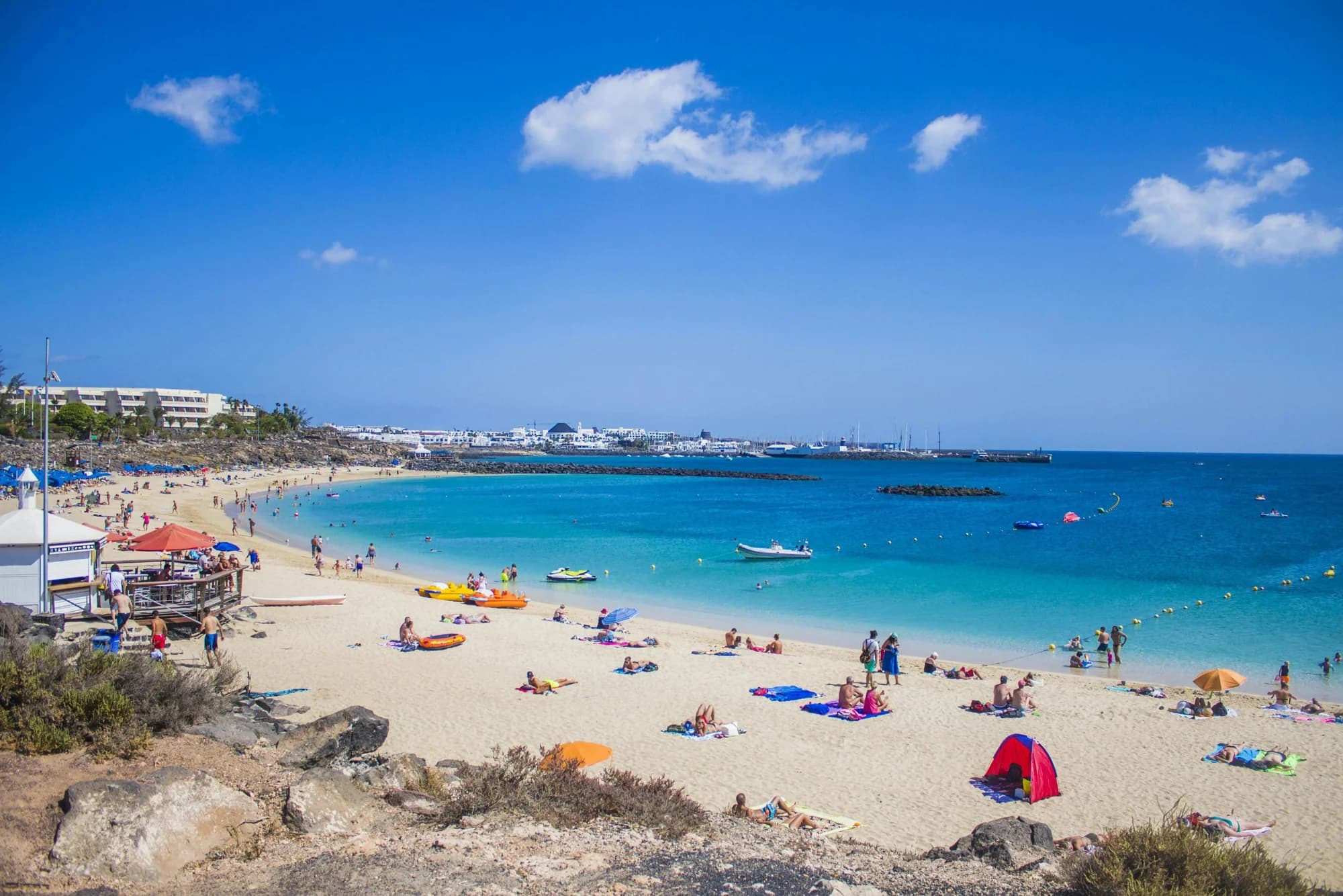 Sandy bay with turquoise water, beachgoers and a marina in the distance